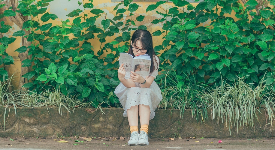 Girl sitting and reading a book in front of green plants.