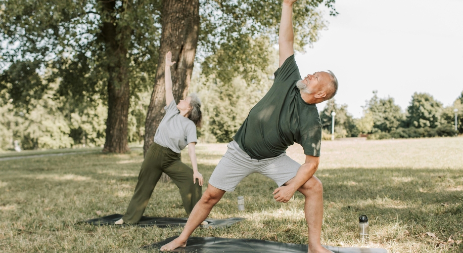 Two people practicing yoga on mats in a park, stretching with one arm raised.