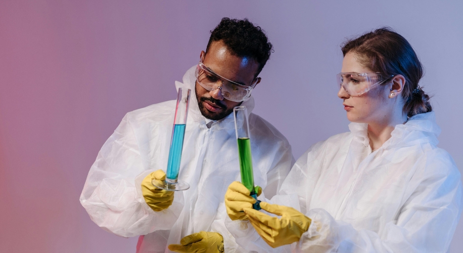 Two scientists examining blue and green liquids in test tubes, wearing protective gear.