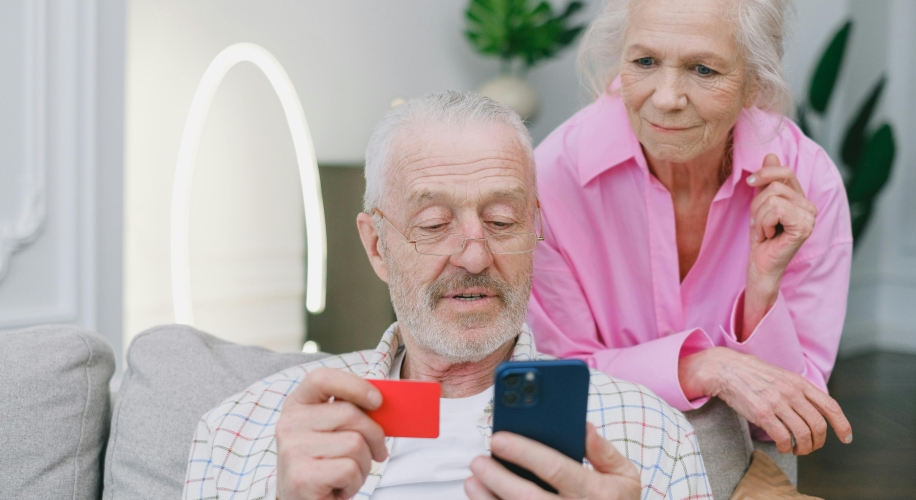 Elderly man holding a red card and looking at a smartphone, with a woman leaning over his shoulder.