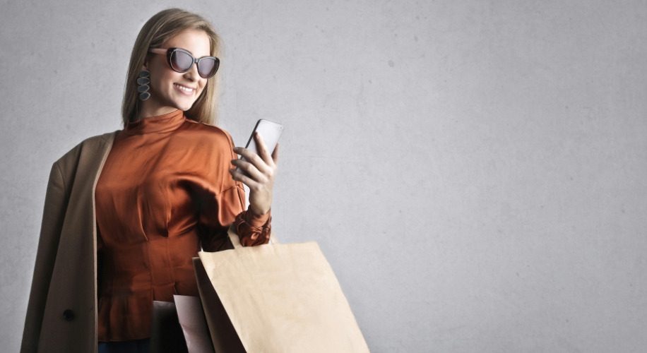 Person holding shopping bags and a smartphone, wearing sunglasses and a brown blouse.