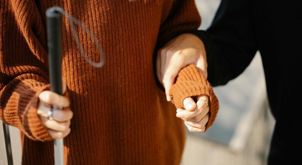 Person holding a white cane, wearing a brown sweater, being guided by another person.