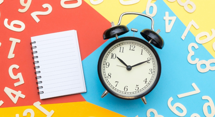 Black analog alarm clock next to an open blank spiral notepad. Background has scattered wooden numbers.