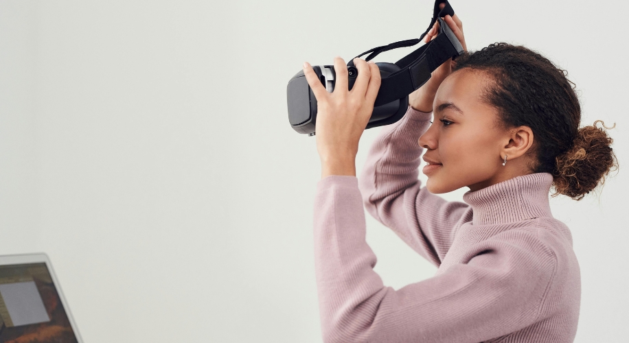 Woman in a pink sweater holding a black VR headset near her face.