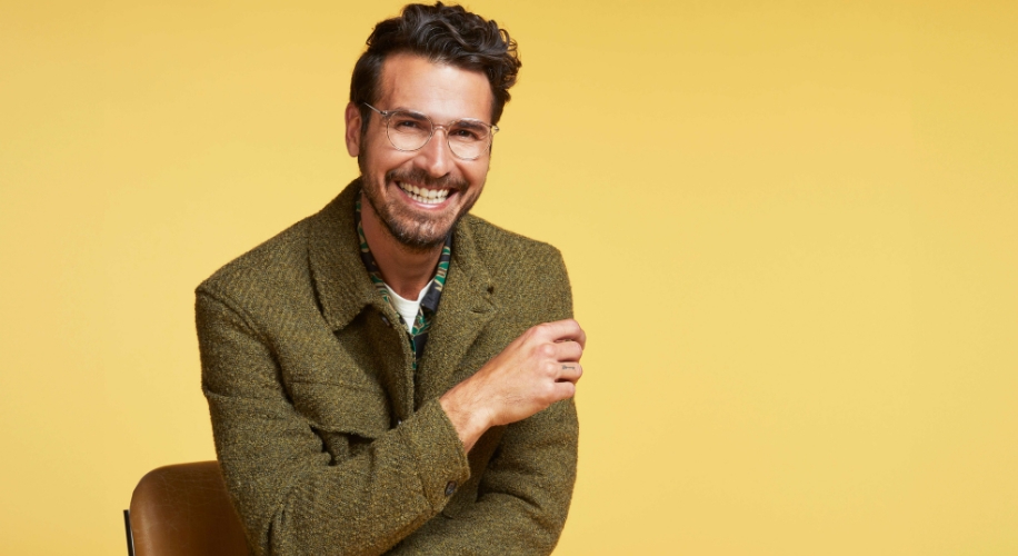 Man wearing a green tweed jacket and clear glasses, smiling against a yellow background.