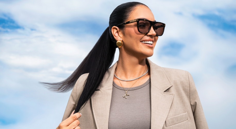 Woman wearing brown sunglasses and a light beige blazer, smiling against a blue sky.