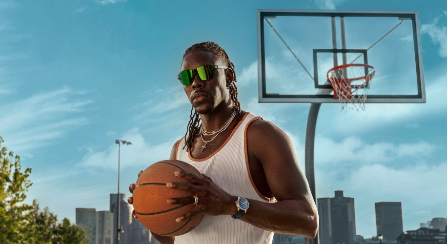 Man wearing reflective sunglasses holds a basketball, standing near a hoop.