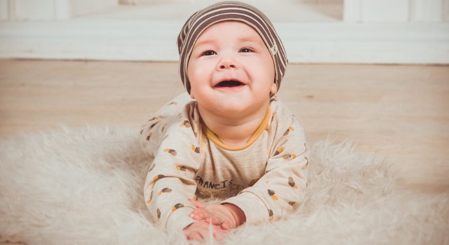 Smiling baby in a striped hat and patterned onesie lying on a fluffy rug.