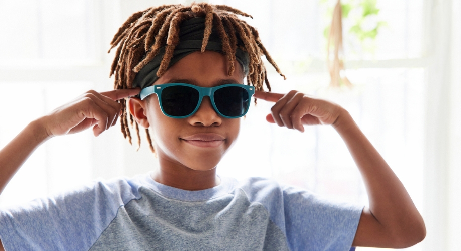 Child wearing blue sunglasses and a gray shirt, pointing at the sunglasses with both hands.