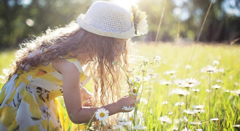 Girl in a white sunhat and floral dress picking daisies in a sunlit field.