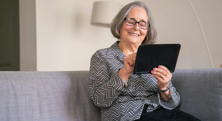 Elderly woman using a tablet, smiling, sitting on a gray couch.